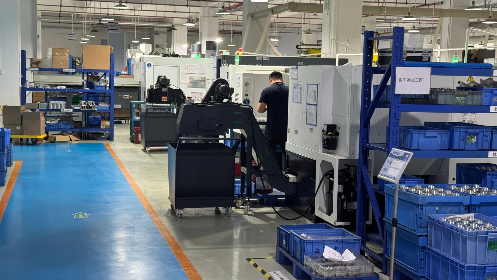 CNC turning workshop with an operator beside a lathe, a marked blue aisle and racks of parts awaiting machining